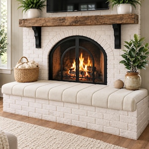 May include: A white brick fireplace hearth featuring a long, cream-colored upholstered bench seat. The fireplace has a black arched metal screen with a fire burning inside, topped by a rustic wooden mantel and two potted green plants.