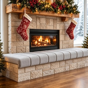 Puede incluir: Una chimenea de piedra con repisa de madera presenta un fuego encendido, dos calcetines navideños rojos con patrones de copos de nieve blancos y un banco tapizado gris integrado en la base de piedra, con una guirnalda verde y bayas.
