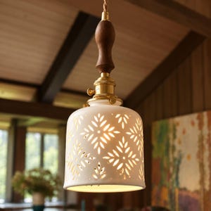 May include: A pendant light featuring a white ceramic shade with a cut-out leaf pattern. The shade is attached to a brass socket and a dark brown wooden handle, suspended by a twisted fabric cord. Light shines through the intricate openings.