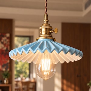 May include: A pendant light featuring a pleated, light blue ceramic shade with a white underside. The fixture includes a brass-toned metal socket with a small side knob and a twisted brown cord, holding a clear, glowing Edison-style light bulb.