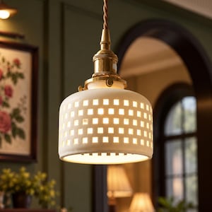 Peut inclure: Une suspension lumineuse dotée d'un abat-jour cylindrique en céramique blanche avec une grille de découpes carrées. L'abat-jour est suspendu par un cordon en tissu marron torsadé et fixé à une douille et une armature en laiton métallique.