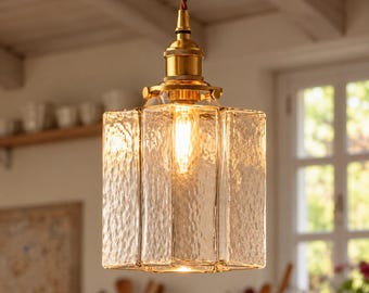 Könnte beinhalten: Eine Pendelleuchte mit einem quadratischen, strukturierten Glasschirm mit gehämmertem Finish. Der Schirm ist an einem braunen, gedrehten Kabel aufgehängt und an einer Fassung sowie Beschlägen aus poliertem Messing befestigt, die eine leuchtende Glühbirne enthalten.