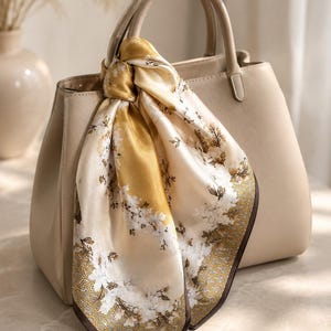 May include: A beige leather handbag with two top handles, adorned with a tied silk scarf featuring a white floral pattern on a gold and cream background, with a dark brown geometric border along the edges of the scarf fabric.