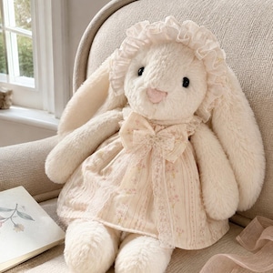 Puede incluir: Un juguete de peluche de conejo color crema con orejas largas, que lleva un gorro a juego con borde con volantes y un vestido crema con un delicado estampado floral rosa, rayas verticales, ribete de encaje y un lazo central.