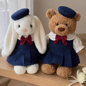 Puede incluir: Un conejo de peluche blanco y un oso de peluche marrón sentados uno al lado del otro. Ambos animales de peluche llevan boinas azul marino a juego, vestidos plisados azul marino con cuellos blancos y lazos de satén rojo oscuro en el cuello.