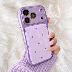 May include: A lavender smartphone case with a glossy finish and a pattern of small, darker purple polka dots. The case features a metallic lavender camera frame with three circular lens cutouts and gold-toned rings around each lens.