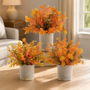 May include: Three artificial plants in cylindrical gray concrete-style pots. Each plant features vibrant orange and yellow foliage with varying leaf shapes, including rounded and needle-like textures, arranged in a dense, bushy display.