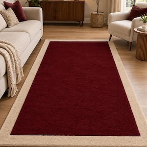 May include: A rectangular area rug featuring a deep burgundy red centre panel surrounded by a wide, solid beige border. The rug has a low-pile texture and is positioned flat on a light-coloured wooden floor, showcasing a clean, two-tone design.