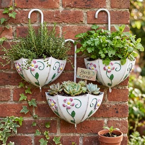 Puede incluir: Tres maceteros de pared de metal blanco con forma de paraguas y ganchos curvos, decorados con flores pintadas en rosa y blanco y enredaderas verdes. Los maceteros contienen hierbas y suculentas, y un letrero de madera dice Herb Corner.