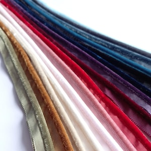 May include: A collection of velvet ribbons arranged in a fan pattern, featuring a spectrum of colors including sage green, golden brown, cream, pale pink, bright red, deep purple, navy blue, and dark teal, each with a soft, textured finish.