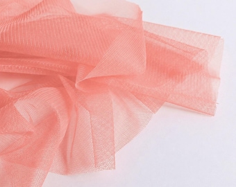 May include: A sheer, lightweight coral-coloured tulle fabric with a fine, open-weave mesh texture. The material is draped in soft, flowing folds, creating layers of translucent, delicate fabric that catch the light across its surface.