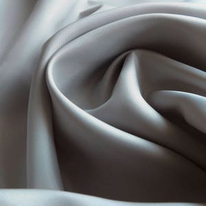 Può includere: Primo piano di un tessuto liscio di colore grigio con una texture morbida e fluida. Il tessuto è drappeggiato e piegato, creando una sensazione di movimento e fluidità.