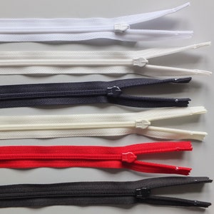 May include: Assortment of five zipper closures in various colors, including white, black, and red. The zippers are laid out horizontally on a gray surface.