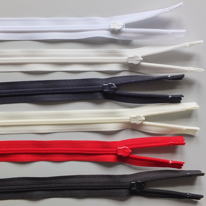 May include: Assortment of five zipper closures in various colors, including white, black, and red. The zippers are laid out horizontally on a gray surface.
