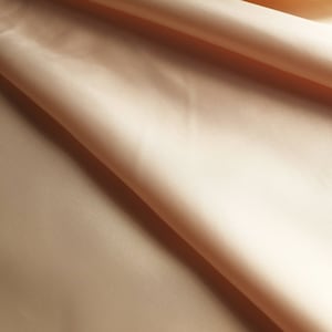 Könnte beinhalten: Nahaufnahme eines glatten, pfirsichfarbenen Stoffes mit weicher, seidenartiger Textur. Der Stoff ist gefaltet und drapiert und bildet eine sanfte Kurve.