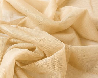 May include: A sheer, light beige fabric with a fine, semi-transparent mesh texture. The material is gathered into soft, flowing folds and drapes, creating a series of gentle curves and shadows that highlight the delicate, lightweight nature of the weave.