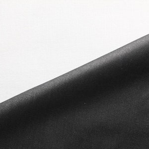 Könnte beinhalten: Nahaufnahme eines schwarzen Stoffes mit glatter, seidenartiger Textur. Der Stoff ist in der Mitte gefaltet und erzeugt eine diagonale Linie über das Bild. Der Hintergrund ist ein weißer Stoff mit leicht strukturierter Oberfläche.