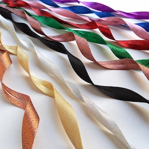 May include: A selection of twisted ribbons in a range of colours, including gold, silver, black, copper, red, green, pink, purple, and blue. The ribbons are arranged diagonally across a white surface. The ribbons are in a variety of metallic and solid finishes.