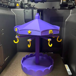 Pode incluir: Um expositor de estilo carrossel roxo impresso em 3D com um pilar central, uma base recortada e um telhado com estrelas amarelas e logotipos do McDonald's. Seis ganchos amarelos pendem do telhado para pendurar itens.