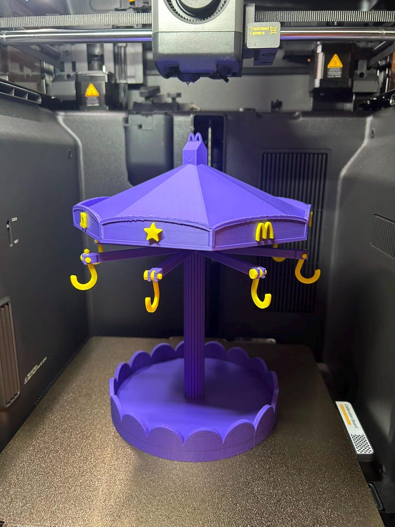 Pode incluir: Um expositor de estilo carrossel roxo impresso em 3D com um pilar central, uma base recortada e um telhado com estrelas amarelas e logotipos do McDonald's. Seis ganchos amarelos pendem do telhado para pendurar itens.