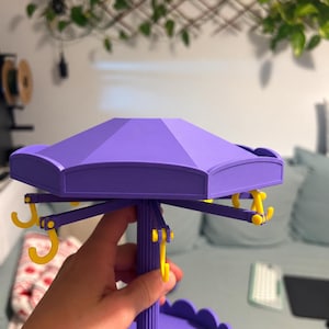Pode incluir: Um organizador de joias estilo carrossel na cor roxa com um teto hexagonal e um pilar central estriado. A estrutura inclui v&aacute;rios ganchos amarelos que se estendem da se&ccedil;&atilde;o superior e uma base circular com borda festonada.