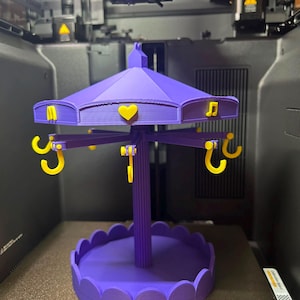 Pode incluir: Um suporte de joias roxo impresso em 3D estilo carrossel com base recortada e pilar central. O telhado apresenta detalhes amarelos, incluindo um cora&ccedil;&atilde;o, uma nota musical e uma letra M. Ganchos amarelos pendem do telhado para armazenamento.