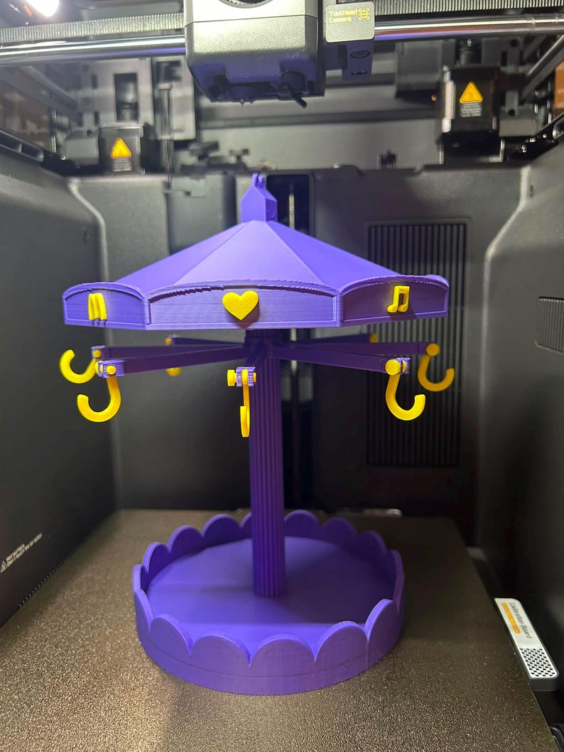 Pode incluir: Um suporte de joias roxo impresso em 3D estilo carrossel com base recortada e pilar central. O telhado apresenta detalhes amarelos, incluindo um cora&ccedil;&atilde;o, uma nota musical e uma letra M. Ganchos amarelos pendem do telhado para armazenamento.