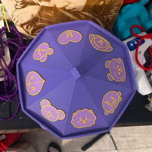 Pode incluir: Uma tampa octogonal roxa com oito &iacute;cones de personagens distintos com contornos amarelos dispostos em um padr&atilde;o circular. Os &iacute;cones incluem v&aacute;rios rostos e formas estilizadas, como um cora&ccedil;&atilde;o, uma nuvem e v&aacute;rias figuras animais em uma superf&iacute;cie roxa.