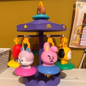 Pode incluir: Um brinquedo de carrossel roxo com v&aacute;rias figuras de personagens penduradas em v&aacute;rias cores, incluindo rosa, amarelo e roxo claro. O topo do carrossel exibe um pequeno personagem marrom e um logotipo amarelo do McDonald's, com estrelas e cora&ccedil;&otilde;es amarelos.