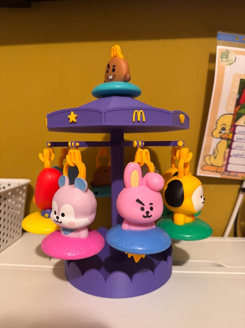 Pode incluir: Um brinquedo de carrossel roxo com v&aacute;rias figuras de personagens penduradas em v&aacute;rias cores, incluindo rosa, amarelo e roxo claro. O topo do carrossel exibe um pequeno personagem marrom e um logotipo amarelo do McDonald's, com estrelas e cora&ccedil;&otilde;es amarelos.