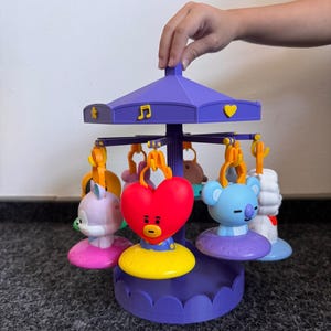 Pode incluir: Um brinquedo de carrossel roxo com v&aacute;rias figuras penduradas, incluindo um cora&ccedil;&atilde;o vermelho, um coala azul-claro e um animal roxo-claro. O teto do carrossel &eacute; decorado com s&iacute;mbolos amarelos como uma estrela, uma nota musical e um cora&ccedil;&atilde;o.