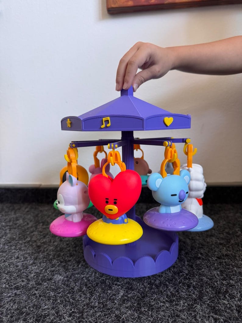 Pode incluir: Um brinquedo de carrossel roxo com v&aacute;rias figuras penduradas, incluindo um cora&ccedil;&atilde;o vermelho, um coala azul-claro e um animal roxo-claro. O teto do carrossel &eacute; decorado com s&iacute;mbolos amarelos como uma estrela, uma nota musical e um cora&ccedil;&atilde;o.