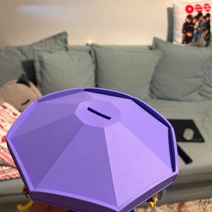 Pode incluir: Um mealheiro octogonal de cor lavanda com um topo plano apresentando uma ranhura retangular central. A estrutura tem um design geom&eacute;trico facetado com pequenos detalhes amarelos vis&iacute;veis ao longo da borda inferior do corpo.