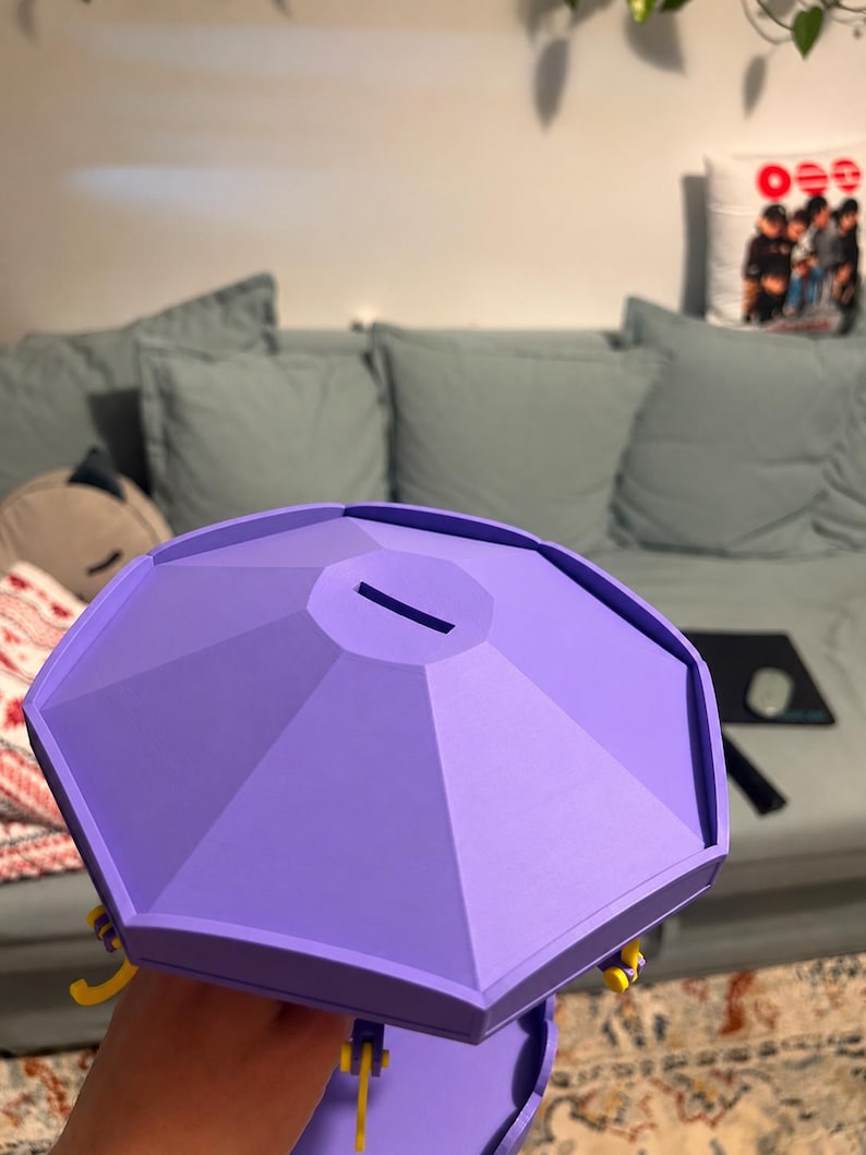 Pode incluir: Um mealheiro octogonal de cor lavanda com um topo plano apresentando uma ranhura retangular central. A estrutura tem um design geom&eacute;trico facetado com pequenos detalhes amarelos vis&iacute;veis ao longo da borda inferior do corpo.