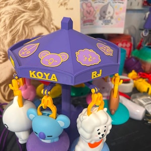 Pode incluir: Um brinquedo de carrossel roxo com figuras de personagens BT21 penduradas em clipes amarelos. O dossel exibe os nomes KOYA e RJ em texto amarelo, juntamente com v&aacute;rios &iacute;cones de personagens. As figuras incluem um coala azul e uma alpaca branca.