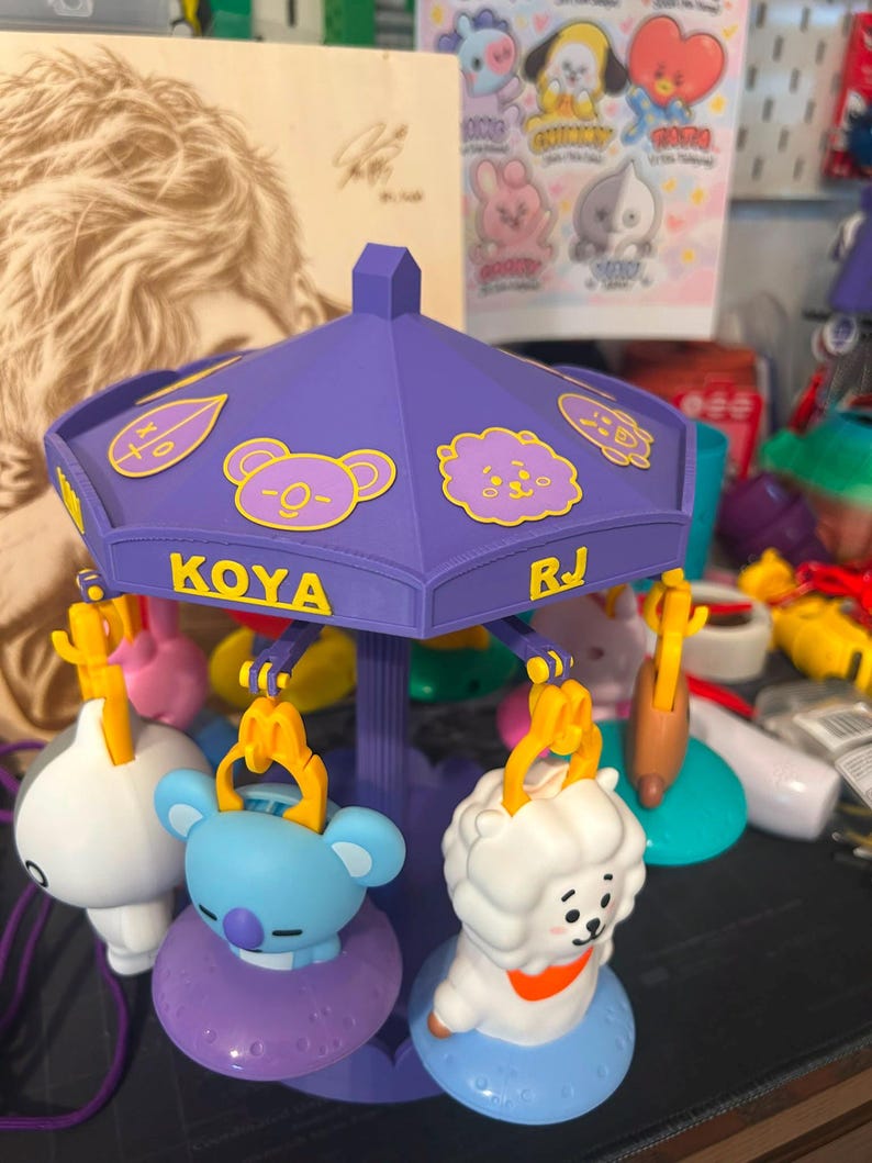Pode incluir: Um brinquedo de carrossel roxo com figuras de personagens BT21 penduradas em clipes amarelos. O dossel exibe os nomes KOYA e RJ em texto amarelo, juntamente com v&aacute;rios &iacute;cones de personagens. As figuras incluem um coala azul e uma alpaca branca.