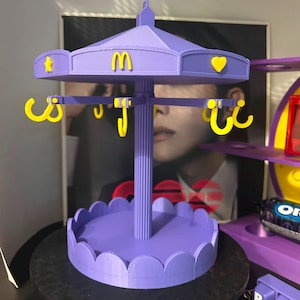 Pode incluir: Um expositor de joias roxo em estilo carrossel com um pilar central, uma base recortada e um topo girat&oacute;rio. O topo &eacute; decorado com &iacute;cones amarelos, incluindo uma estrela, um cora&ccedil;&atilde;o e o logotipo dos arcos dourados do McDonald's.