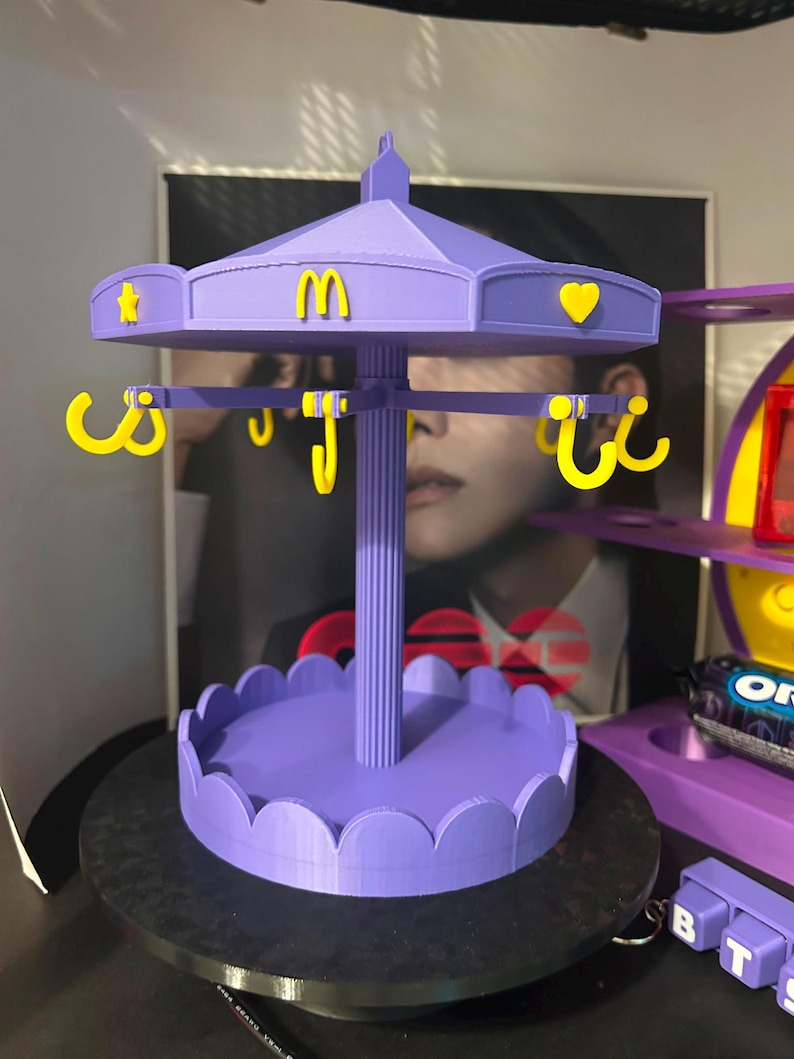 Pode incluir: Um expositor de joias roxo em estilo carrossel com um pilar central, uma base recortada e um topo girat&oacute;rio. O topo &eacute; decorado com &iacute;cones amarelos, incluindo uma estrela, um cora&ccedil;&atilde;o e o logotipo dos arcos dourados do McDonald's.
