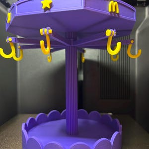 Pode incluir: Um expositor girat&oacute;rio roxo impresso em 3D com um pilar central, uma base festonada e um n&iacute;vel superior com ganchos amarelos. O n&iacute;vel superior &eacute; decorado com estrelas amarelas e a letra M. Estrutura projetada para pendurar objetos.