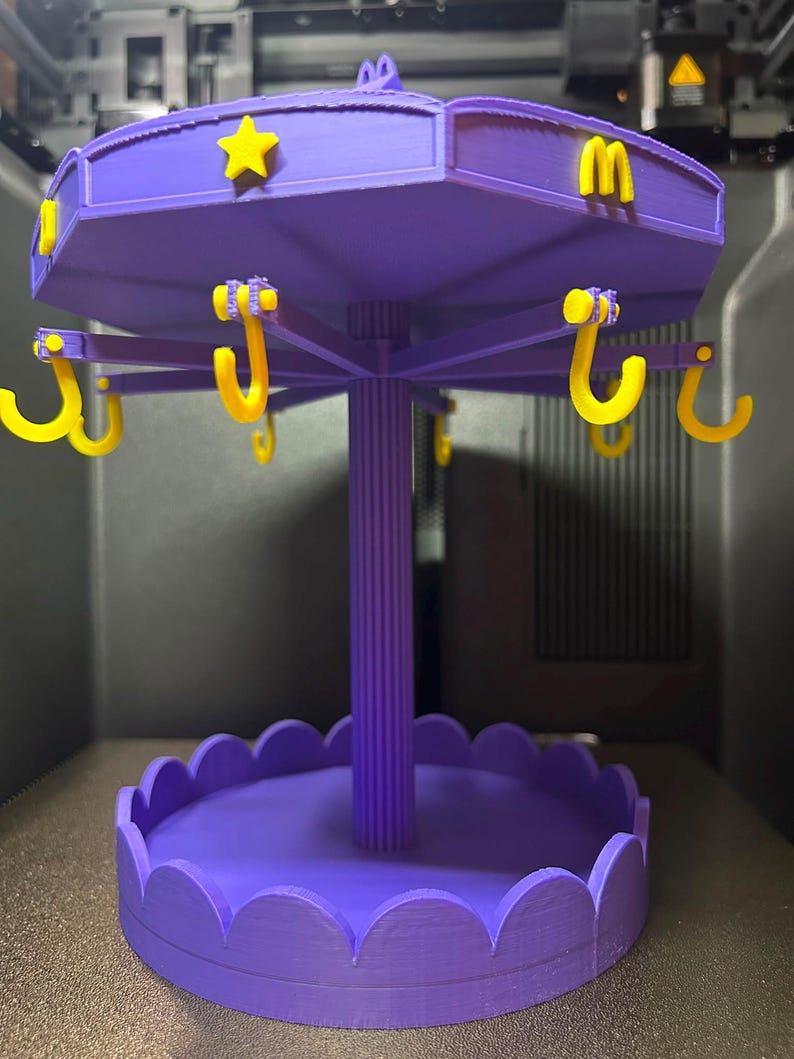 Pode incluir: Um expositor girat&oacute;rio roxo impresso em 3D com um pilar central, uma base festonada e um n&iacute;vel superior com ganchos amarelos. O n&iacute;vel superior &eacute; decorado com estrelas amarelas e a letra M. Estrutura projetada para pendurar objetos.