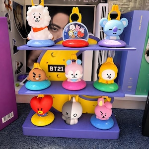Pode incluir: Um expositor roxo de três níveis em forma de meia-lua com o texto BT21, contendo nove pequenas figuras coloridas de personagens. As figuras incluem uma alpaca branca, um coala azul, um cão castanho e uma personagem de coração vermelho.