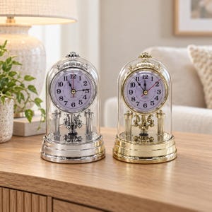 May include: Two mantel clocks with glass domes, one with a silver-tone base and one with a gold-tone base. Each clock features a white face with black Arabic numerals, black hour and minute hands, and a red second hand, supported by pillars.
