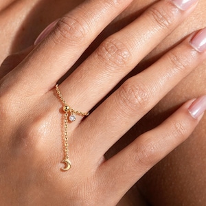 May include: A gold chain ring featuring a delicate dangling charm in the shape of a crescent moon and a small, round, clear gemstone set in a gold bezel. The ring band is constructed from fine gold chain links with a small gold bead accent.