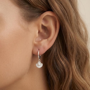 May include: A silver hoop earring featuring a small, dangling seashell charm. The seashell charm is crafted from silver and has a tiny, round white pearl centered at the base of the shell. The earring is designed with a simple, polished silver hoop.