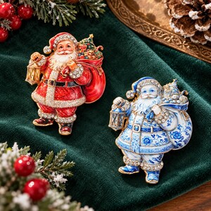 May include: Two Santa Claus brooches featuring clear crystal accents. One Santa wears a red suit with gold trim, while the other wears a blue and white floral patterned suit. Both figures carry a sack over their shoulder and hold a lantern.