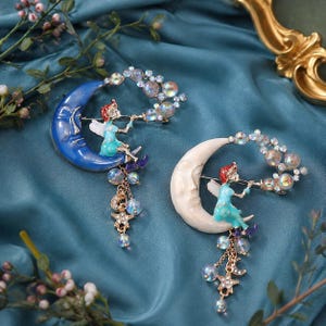 May include: Two crescent moon brooches featuring a seated fairy figure playing a flute. One moon is deep blue, the other is cream-colored. Both designs include iridescent crystal accents, dangling gold-tone chains, small star charms, and moon charms.