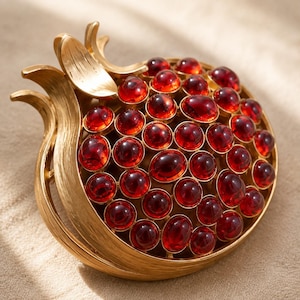 May include: A gold-toned brooch shaped like a pomegranate, featuring a brushed metal finish on the outer structure and numerous round, deep red cabochon stones set within the interior to resemble the fruit's seeds.