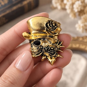 May include: A gold-toned metal brooch featuring a skull wearing a top hat, accented with black enamel details and sculpted roses. The design includes a prominent top hat, a skull with dark eye sockets, and floral elements integrated into the side.