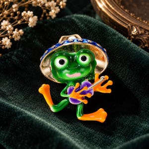 Könnte beinhalten: Eine Metallbrosche in Form eines grünen Frosches mit einem blau-goldenen Hut. Der Frosch hat große weiße Augen mit schwarzen Pupillen, einen rosa Mund und orangefarbene Füße. Er hält ein violett-oranges Instrument mit einem silbernen Hals.