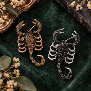 May include: Two scorpion-shaped brooches. One brooch is gold-toned with brown crystal accents covering the body and claws. The second brooch is silver-toned with black crystal accents covering the body and claws. Both feature a pin mechanism.